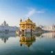 A hyper-realistic photograph of the Golden Temple (Harmandir Sahib) in Amritsar at sunrise. The golden architecture is perfectly centered and reflected in the calm Amrit Sarovar water under a clear morning sky.