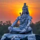 Meditating Lord Shiva statue seated in Padmasana on a tiger skin pedestal, with a glowing orange sunset sky in the background, symbolizing peace, spirituality, and divine power.
